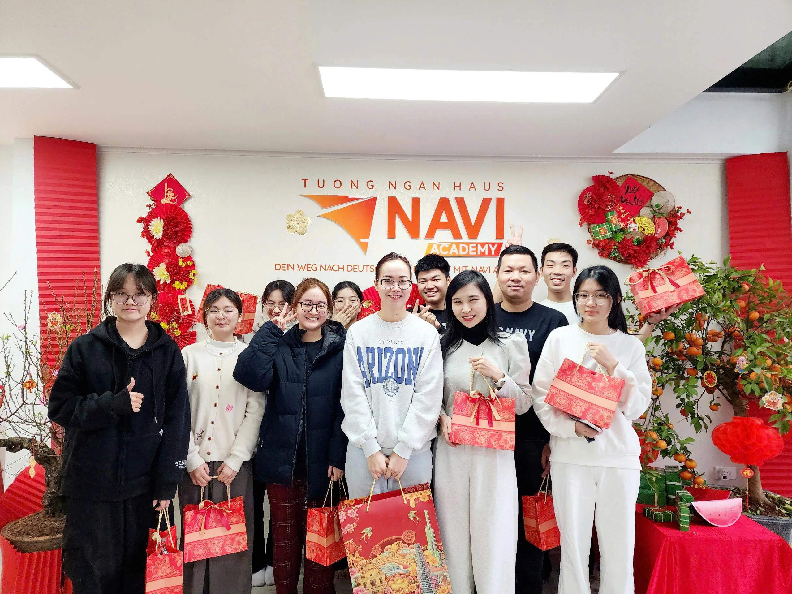 Học viên và giáo viên NAVI Academy chụp ảnh chung, tay cầm túi quà Tết trước bảng hiệu trung tâm.